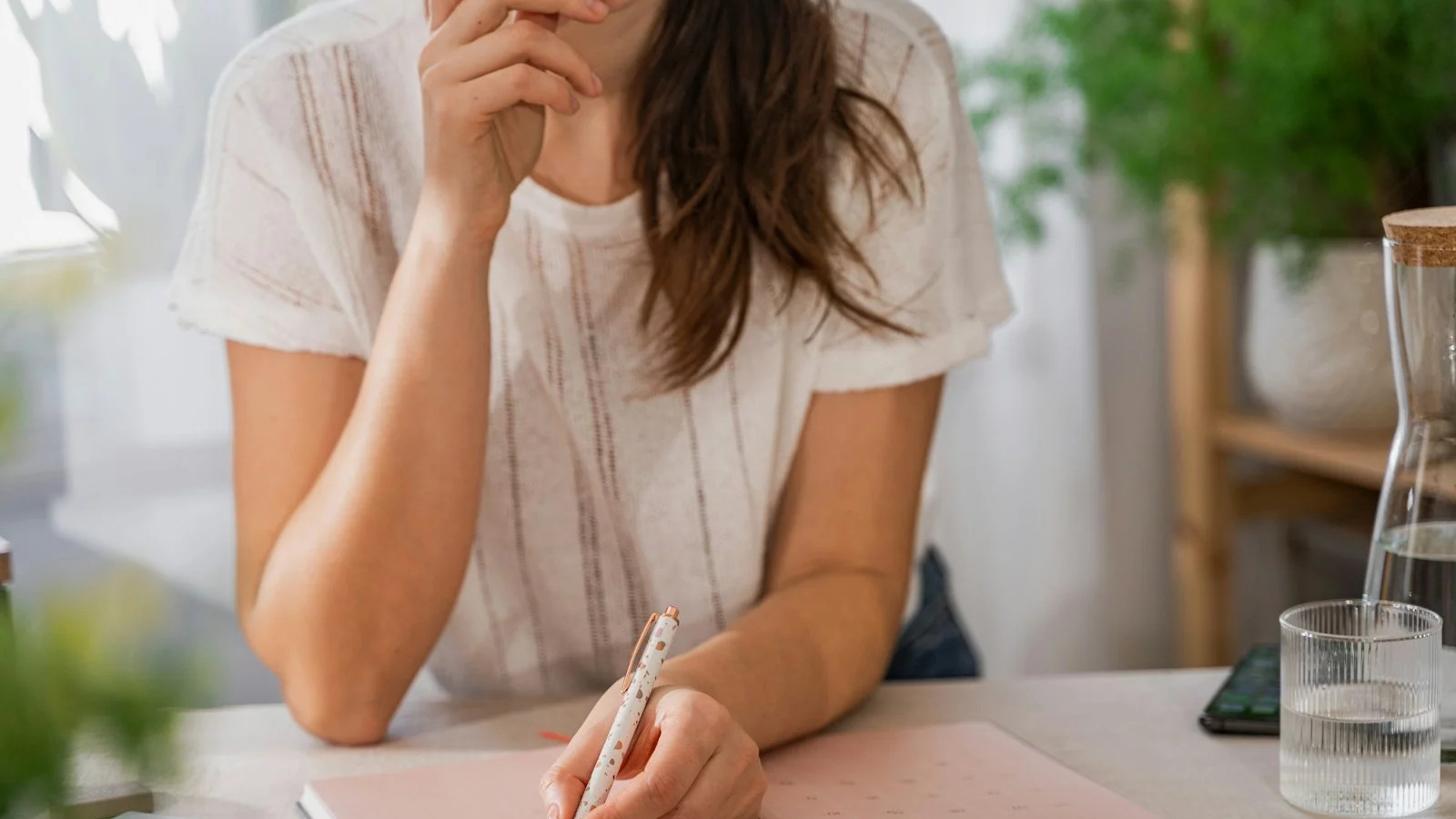 Femme réfléchissant, tenant un stylo, fond végétal, ambiance lumineuse.